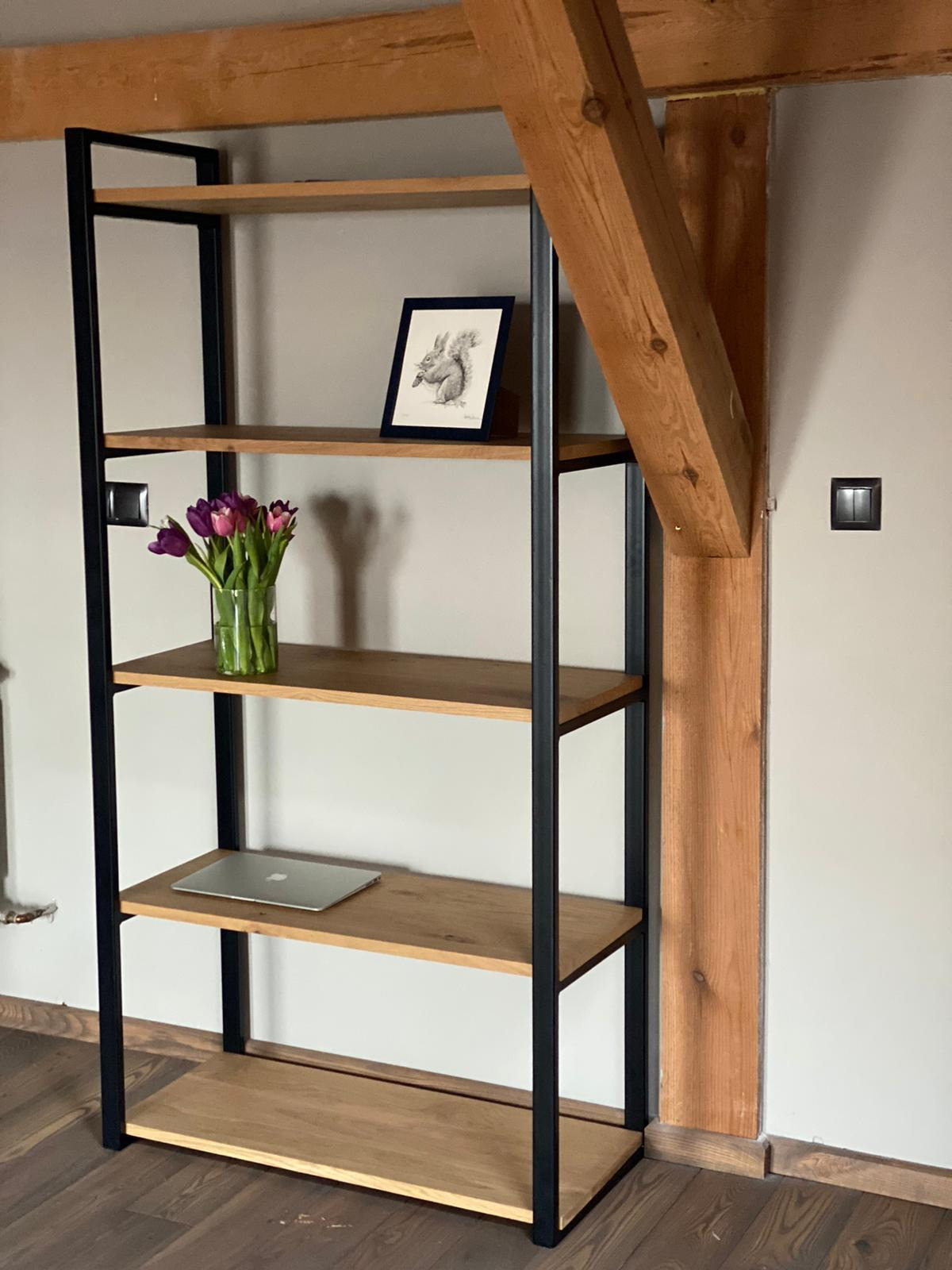 Regał z drewnianymi półkami i czarną metalową ramą, na półkach wazon z tulipanami, ramka ze szkicem wiewiórki i laptop, na tle drewniany element konstrukcyjny i szara ściana.