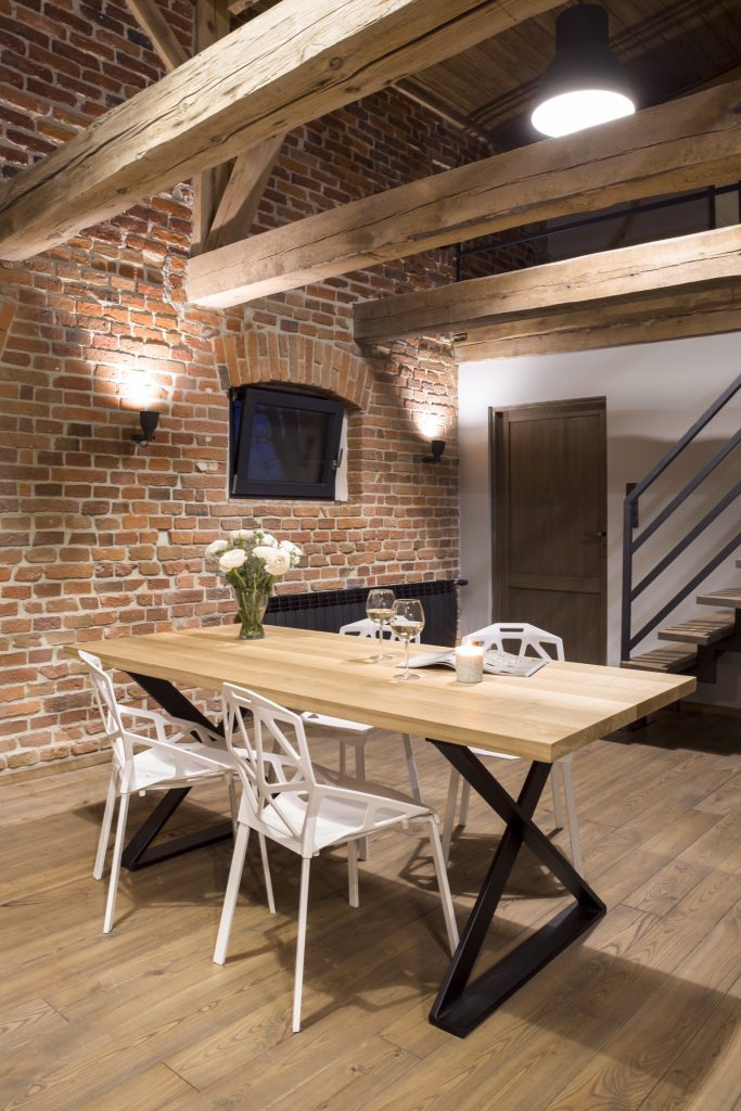 Stół z jasnego drewna z czarnymi, metalowymi nogami w kształcie X, otoczony białymi krzesłami, w rustykalnym wnętrzu z ceglaną ścianą i drewnianymi belkami stropowymi w Pleszewie.