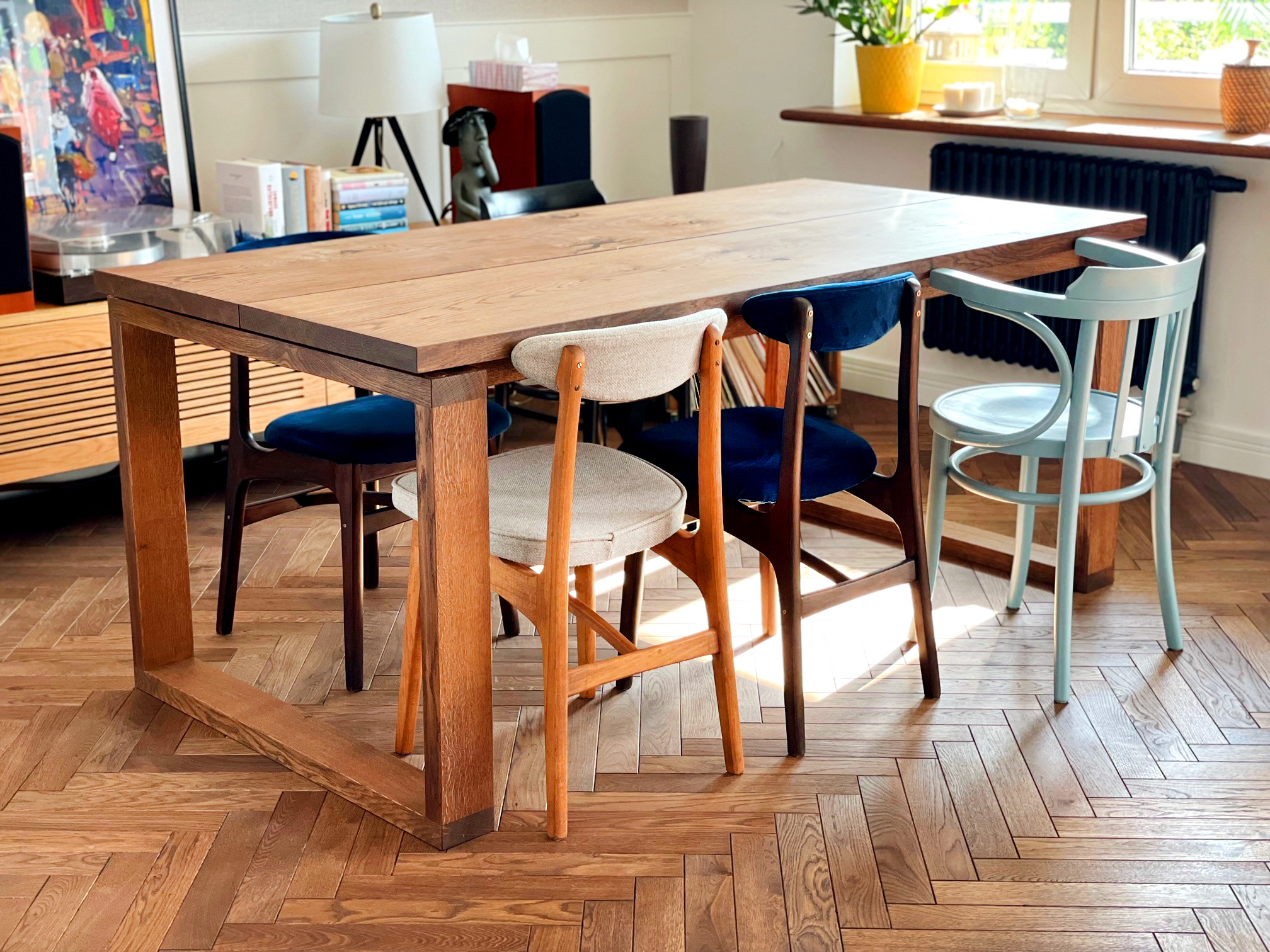 Stół z litego drewna z nogami w kształcie ramy, otoczony krzesłami w różnych kolorach i stylach na parkiecie w jodełkę.