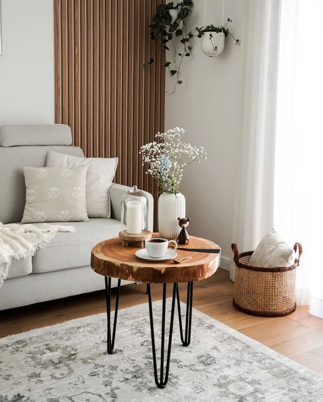 Stolik kawowy z blatem z plastra drewna na metalowych nogach w salonie z szarą sofą, dywanem w kwiaty i drewnianymi panelami ściennymi.