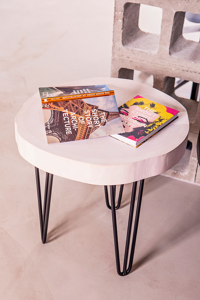 Okrągły stolik kawowy z blatem z jasnego drewna i czarnymi, metalowymi nóżkami typu hairpin, na blacie leżą dwie książki o architekturze i sztuce współczesnej, w tle betonowy ażur.