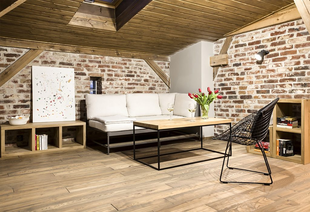 Jasne wnętrze poddasza z drewnianym sufitem i ceglaną ścianą, wyposażone w białą sofę, drewniany stolik kawowy, ażurowe krzesło i regał z książkami.