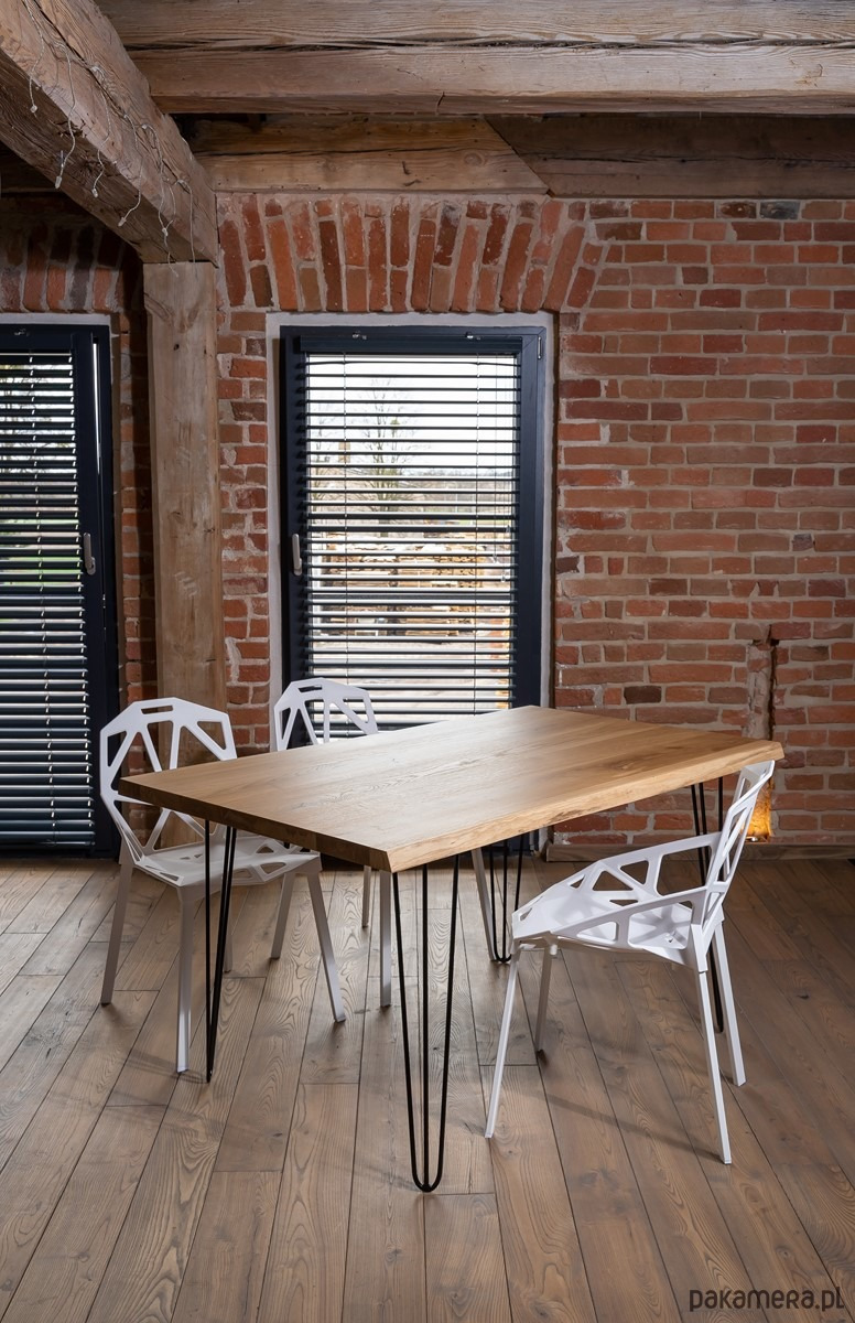 Stół z blatem z jasnego drewna na czarnych, metalowych nogach typu hairpin, otoczony białymi krzesłami o ażurowej konstrukcji, na tle ściany z czerwonej cegły i drewnianych belek stropowych.