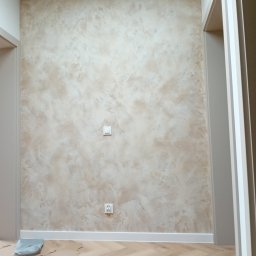 Home serwis-Kafelkowanie - Ściana z dekoracyjną, beżowo-brązową fakturą w mieszkaniu w Ozimku, widoczne gniazdko elektryczne i włącznik światła, zabezpieczona podłoga z panelami ułożonymi w jodełkę.