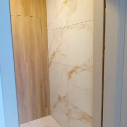 Ściana wyłożona wielkoformatowymi płytkami imitującymi marmur w odcieniach bieli i złota, sąsiadująca z drewnopodobną okładziną ściany. Widoczny fragment podłogi z identycznych płytek.