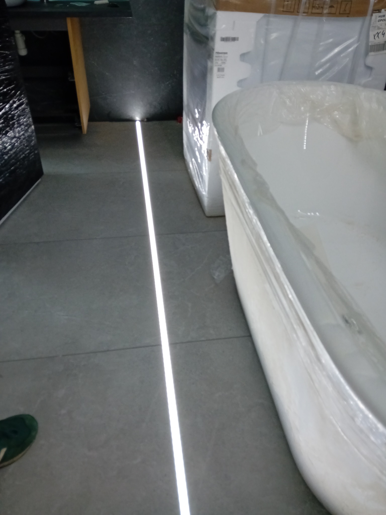 Nowa wanna w folii ochronnej stoi na szarych płytkach podłogowych z oświetleniem liniowym LED wzdłuż ściany, obok zapakowanych pudeł i fragmentu buta.