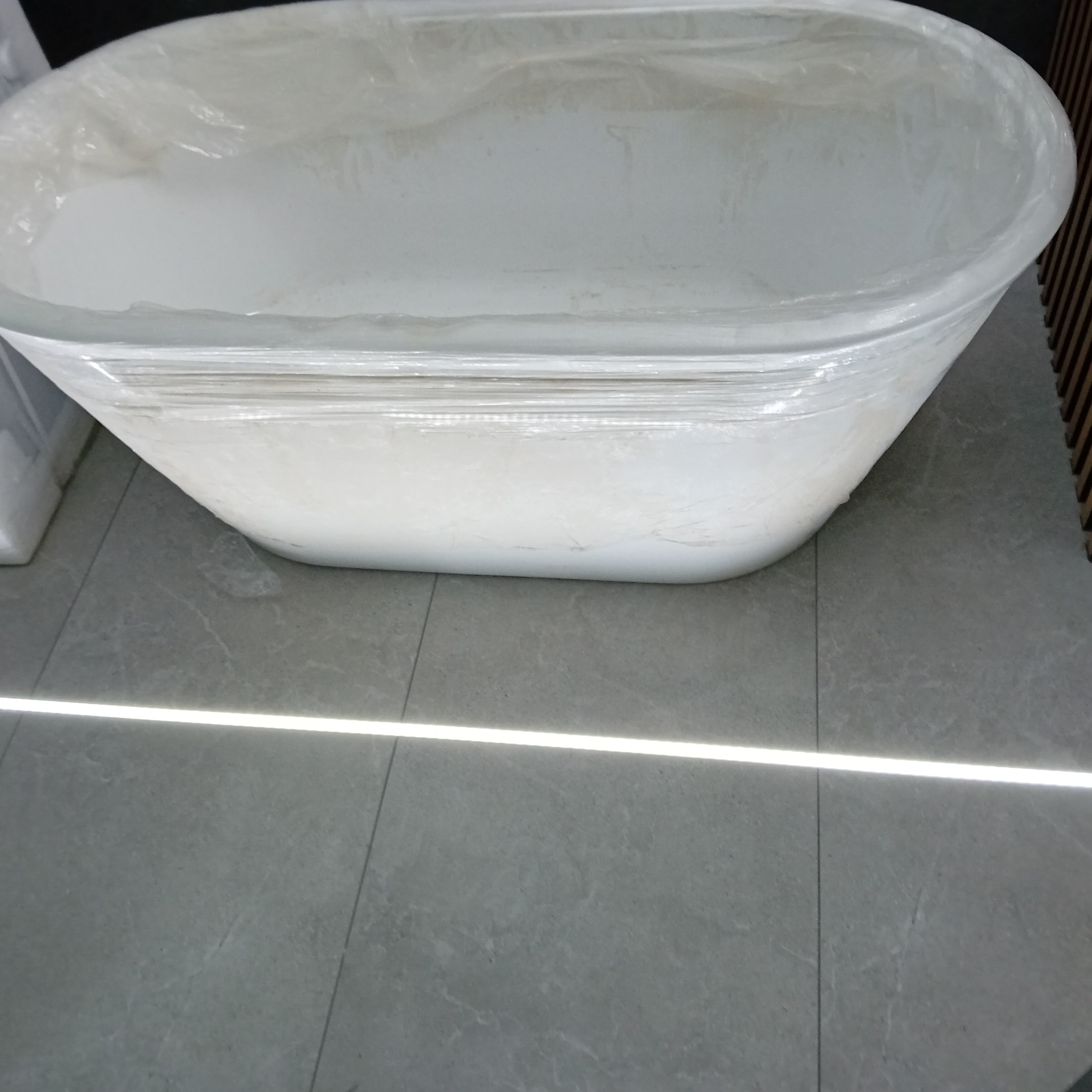 Nowa, owalna wanna wolnostojąca, zabezpieczona folią ochronną, ustawiona na szarych, matowych płytkach podłogowych z delikatnym wzorem.