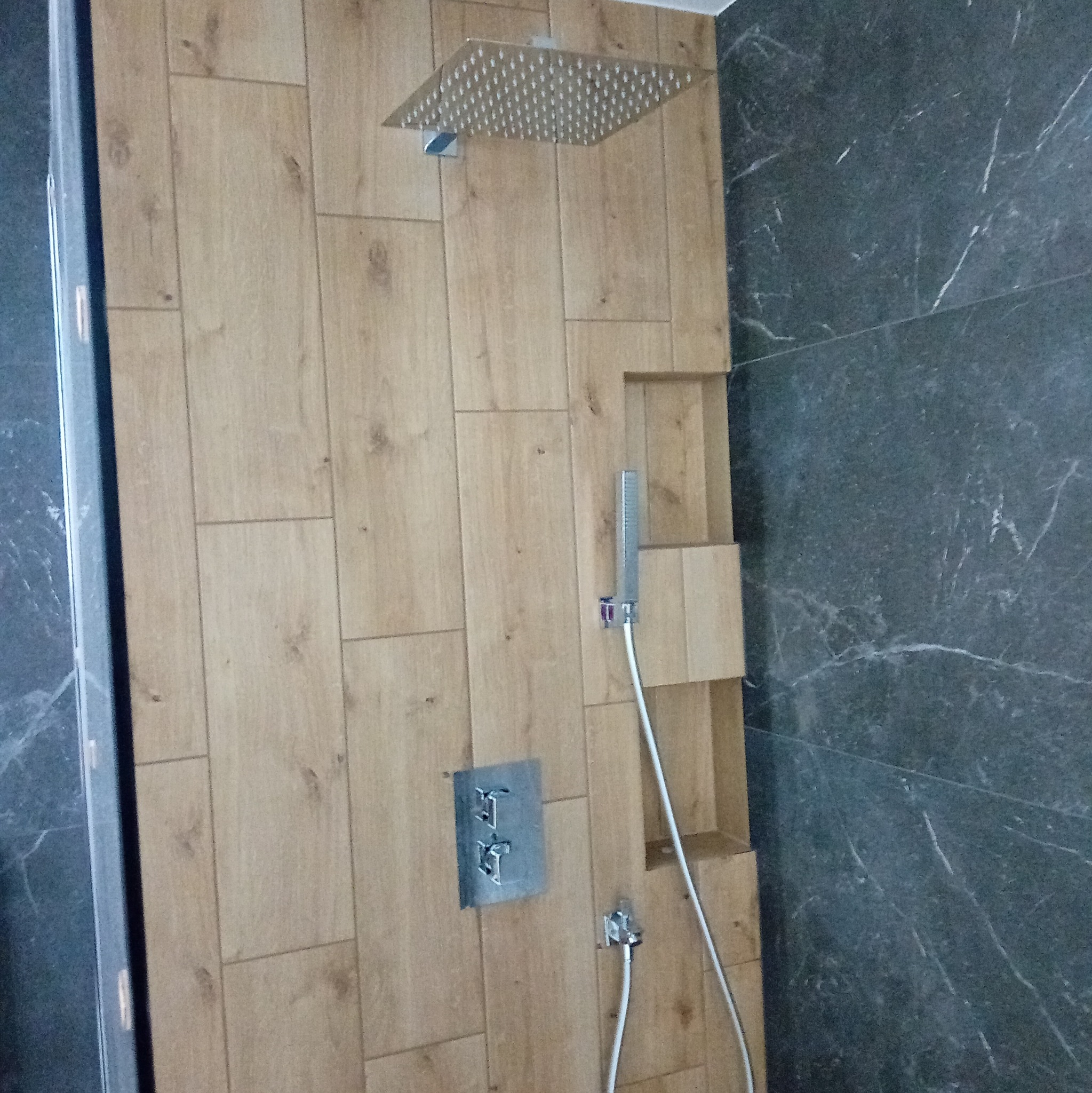 Nowoczesna kabina prysznicowa z drewnopodobnymi płytkami i marmurową ścianą, wyposażona w deszczownicę i słuchawkę prysznicową, z wnęką na kosmetyki.