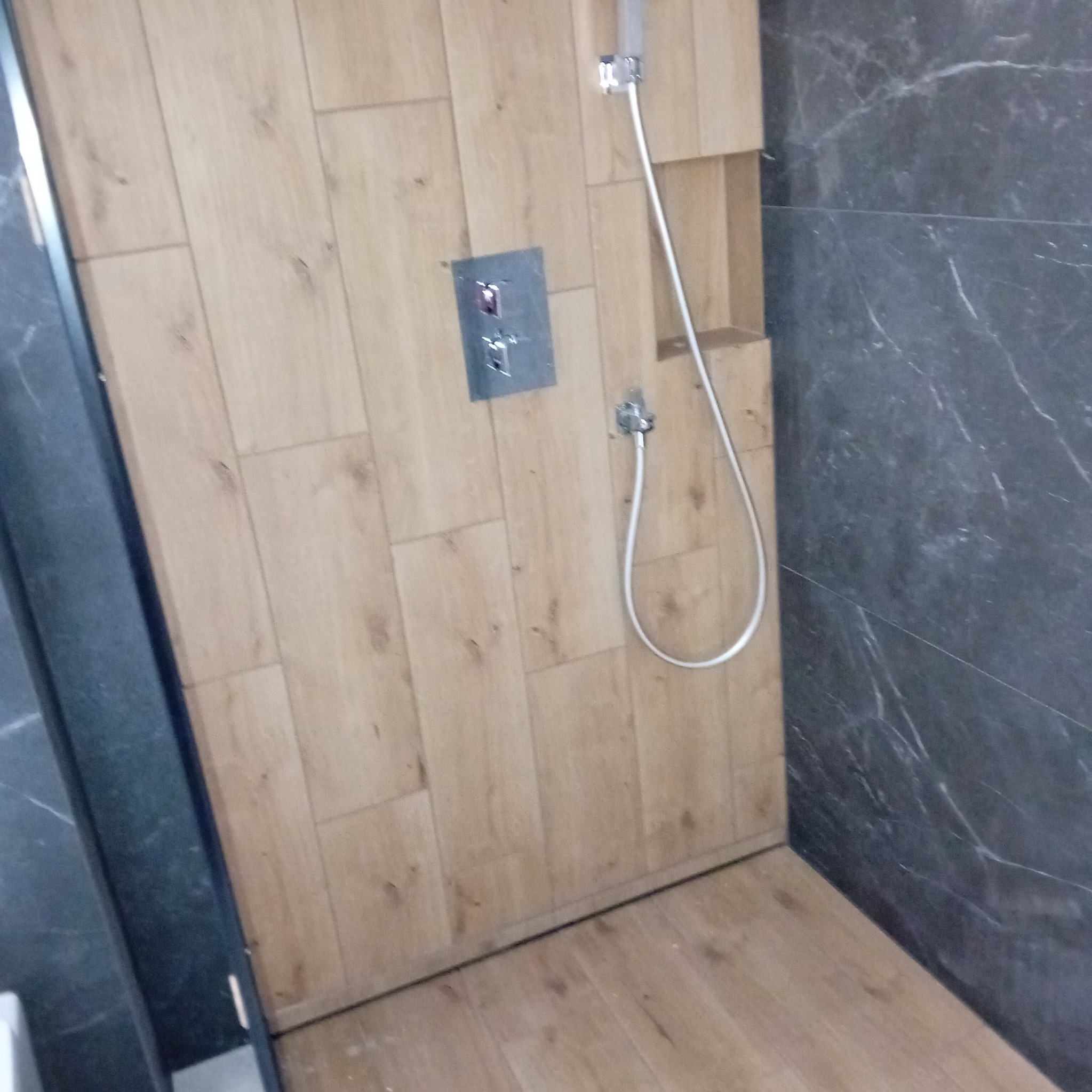 Wykończona kabina prysznicowa z drewnopodobnymi płytkami na ścianach i podłodze, kontrastująca z ciemnoszarym marmurem na jednej ze ścian. Widoczna bateria prysznicowa i wnęka w ścianie.