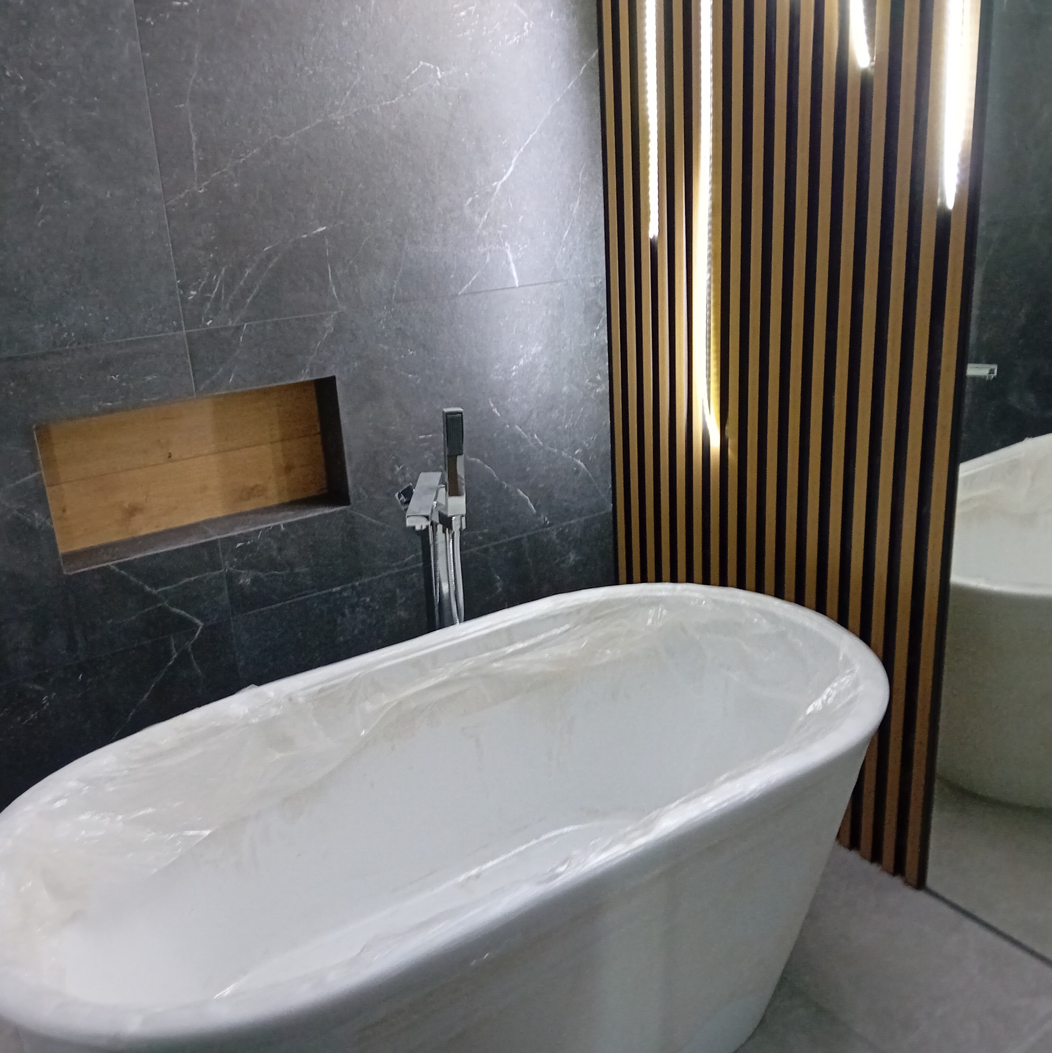 Nowoczesna łazienka z wolnostojącą wanną owiniętą folią ochronną, szare płytki imitujące marmur z wnęką i chromowana bateria.