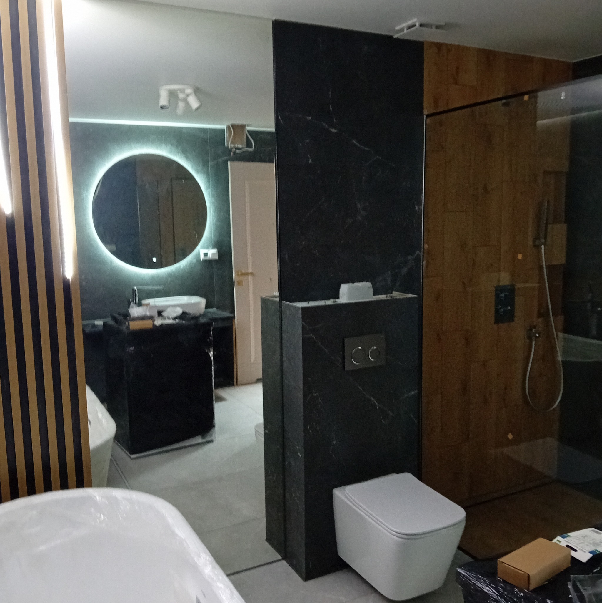 Nowoczesna łazienka w trakcie wykańczania, z podświetlanym lustrem, czarnymi marmurkowymi płytkami i drewnianą kabiną prysznicową.