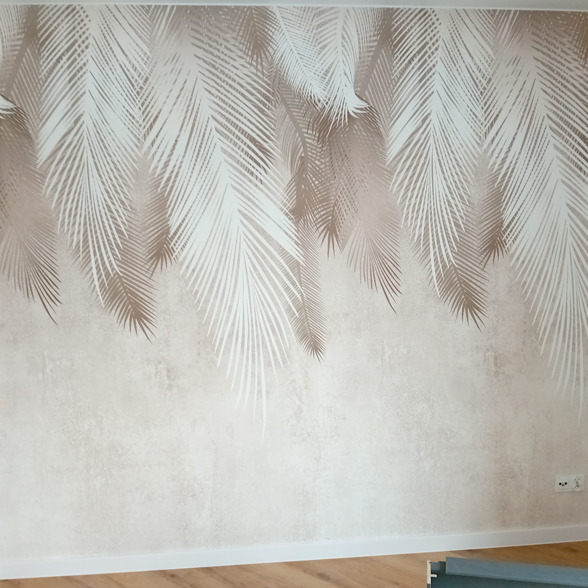 Ściana z beżową tapetą w motywy białych i beżowych liści palmowych, widoczny fragment drewnianej podłogi i listwa przypodłogowa.