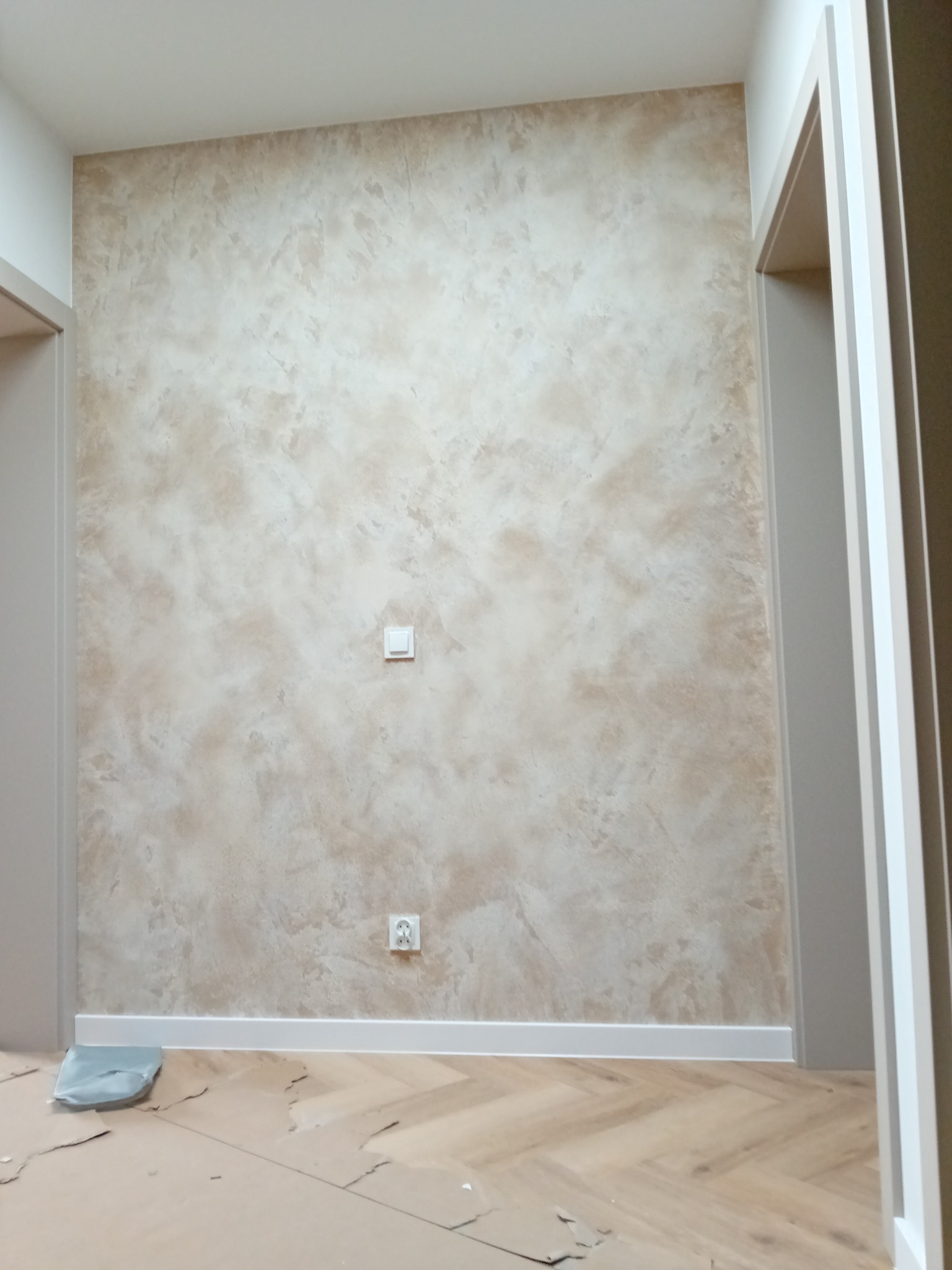 Ściana z dekoracyjną, beżowo-brązową fakturą w mieszkaniu w Ozimku, widoczne gniazdko elektryczne i włącznik światła, zabezpieczona podłoga z panelami ułożonymi w jodełkę.