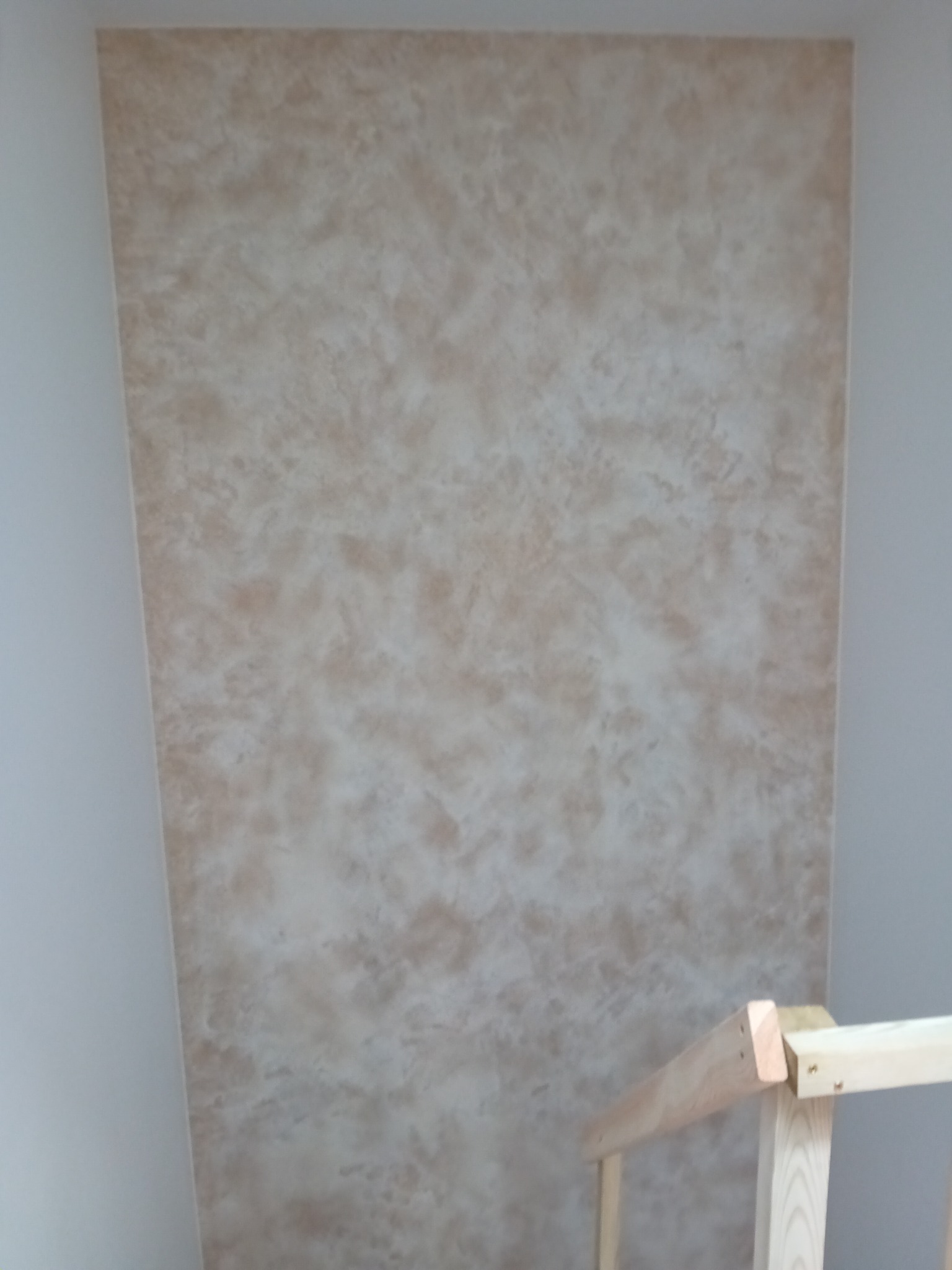 Beżowa ściana z efektem dekoracyjnym, imitującym marmur, widoczny fragment drewnianej barierki w dolnym rogu.