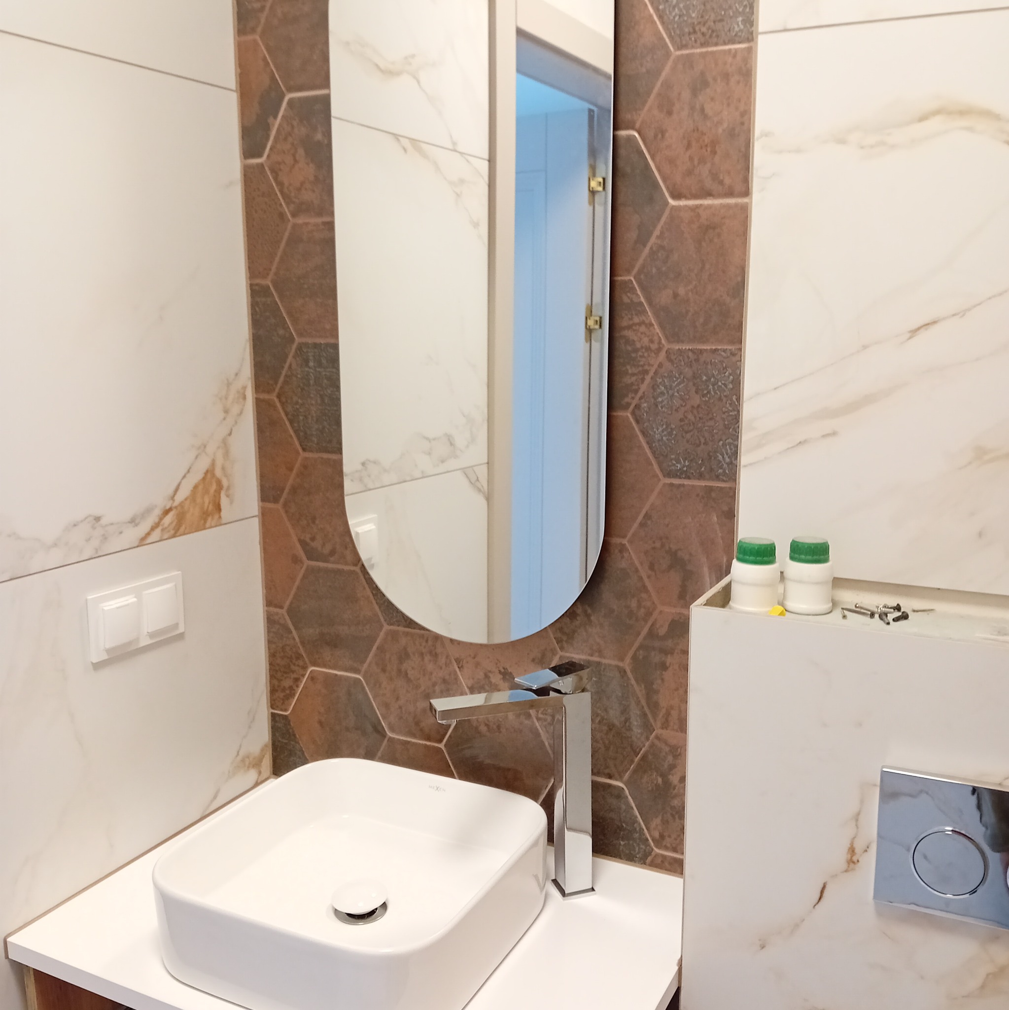 Umywalka nablatowa z chromowaną baterią, owalne lustro, płytki imitujące marmur i heksagonalne płytki w odcieniach brązu, dwie butelki z zielonymi nakrętkami i drobne elementy montażowe na półce.