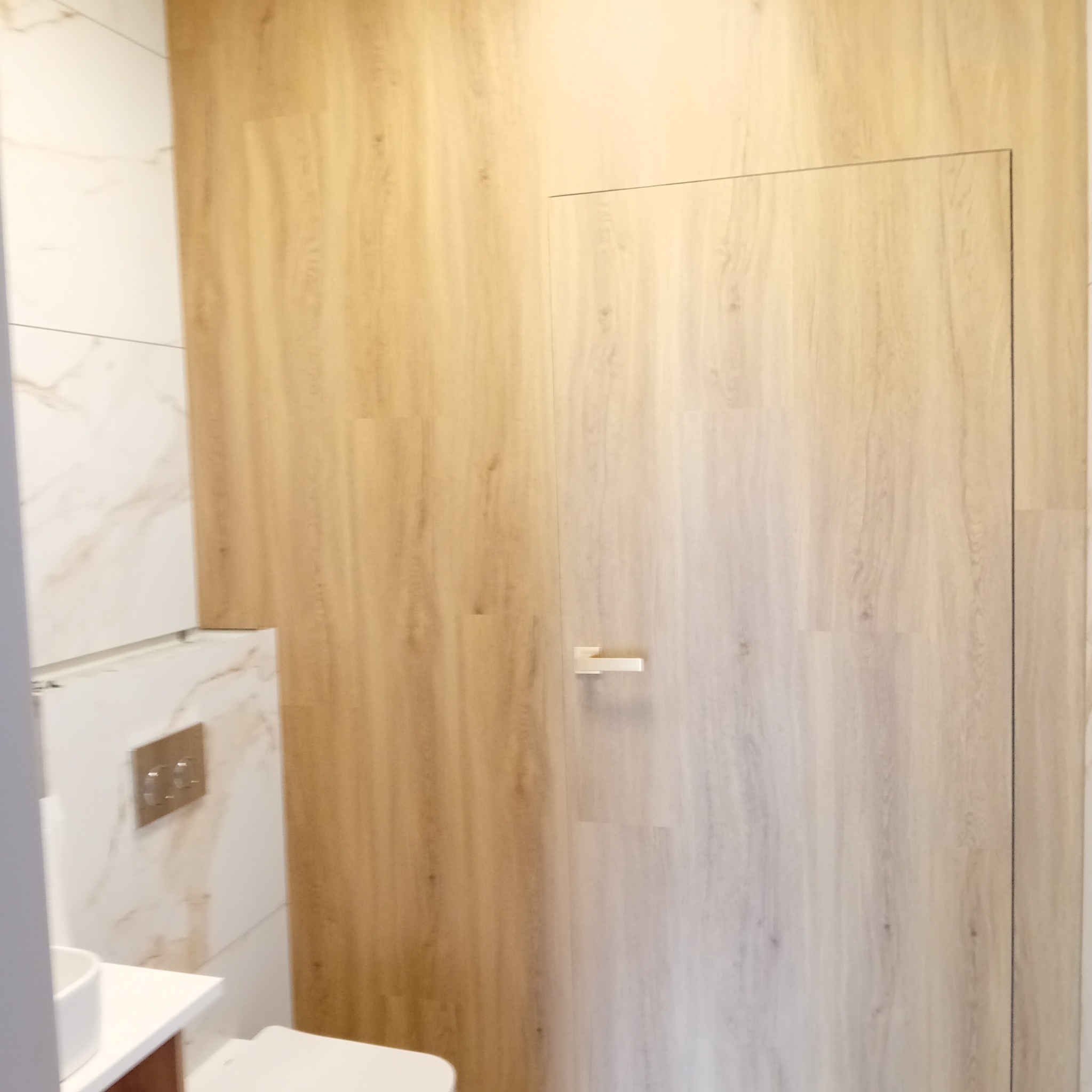 Nowoczesna łazienka z ukrytymi drzwiami wykończonymi panelami ściennymi imitującymi drewno, dopasowanymi kolorystycznie do reszty ścian, obok marmurowe płytki i podwieszana toaleta.