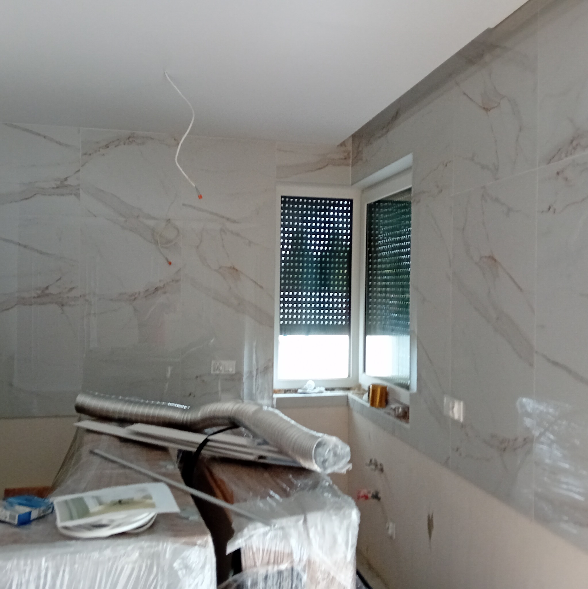 Wnętrze łazienki w trakcie prac wykończeniowych: marmurowe płytki na ścianach, okno z roletami, widoczne przewody elektryczne i materiały budowlane zabezpieczone folią.
