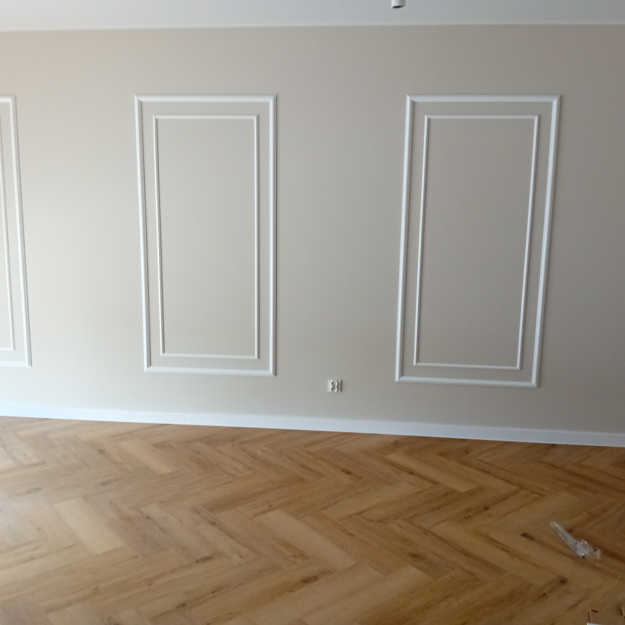 Beżowa ściana z białymi, prostokątnymi sztukateriami i drewniana podłoga ułożona w jodełkę.