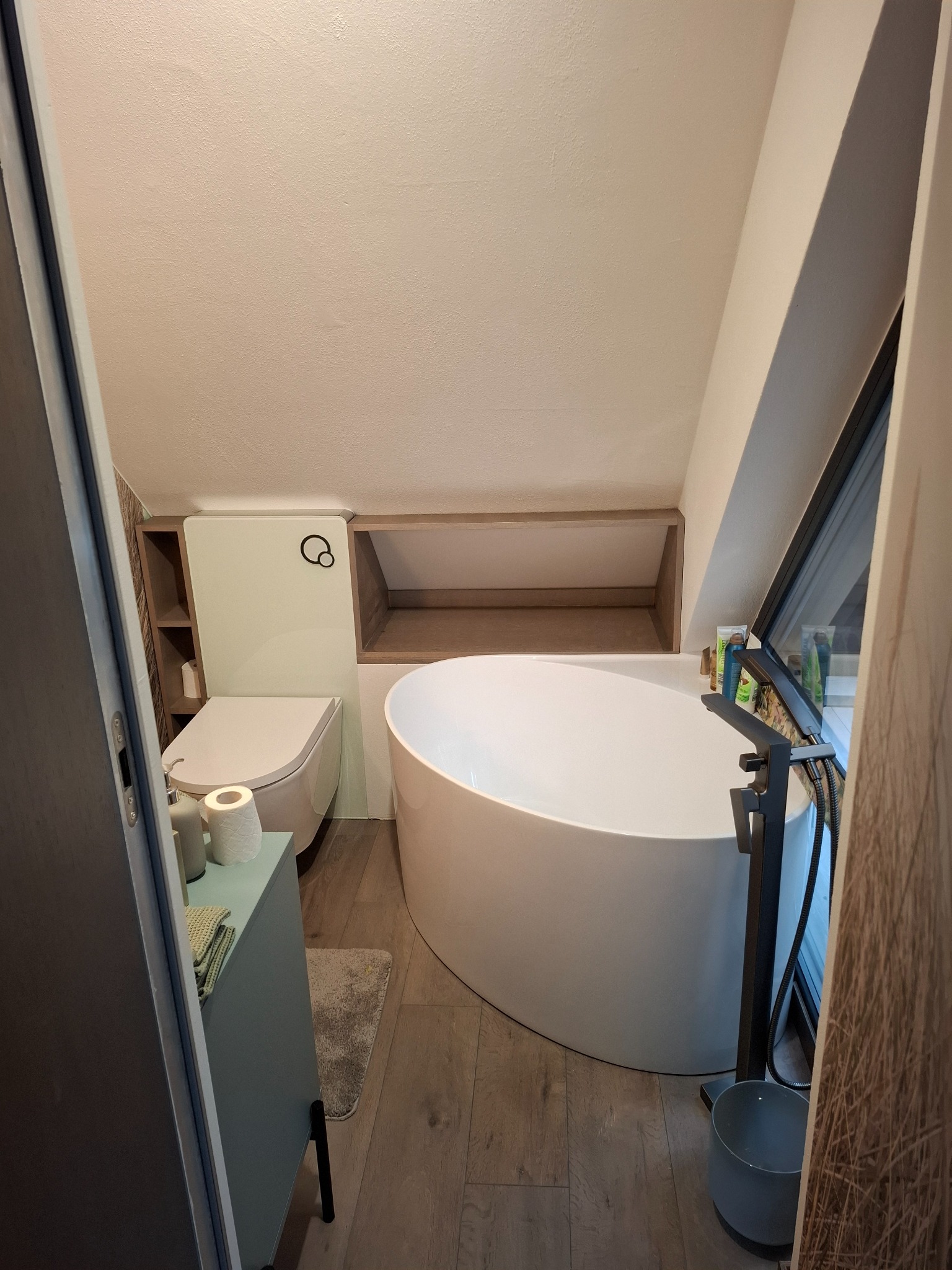 Nowoczesna łazienka na poddaszu z wolnostojącą wanną, podwieszaną toaletą i szafką w odcieniach szarości. Uwagę zwraca okno dachowe i drewniane wykończenie.