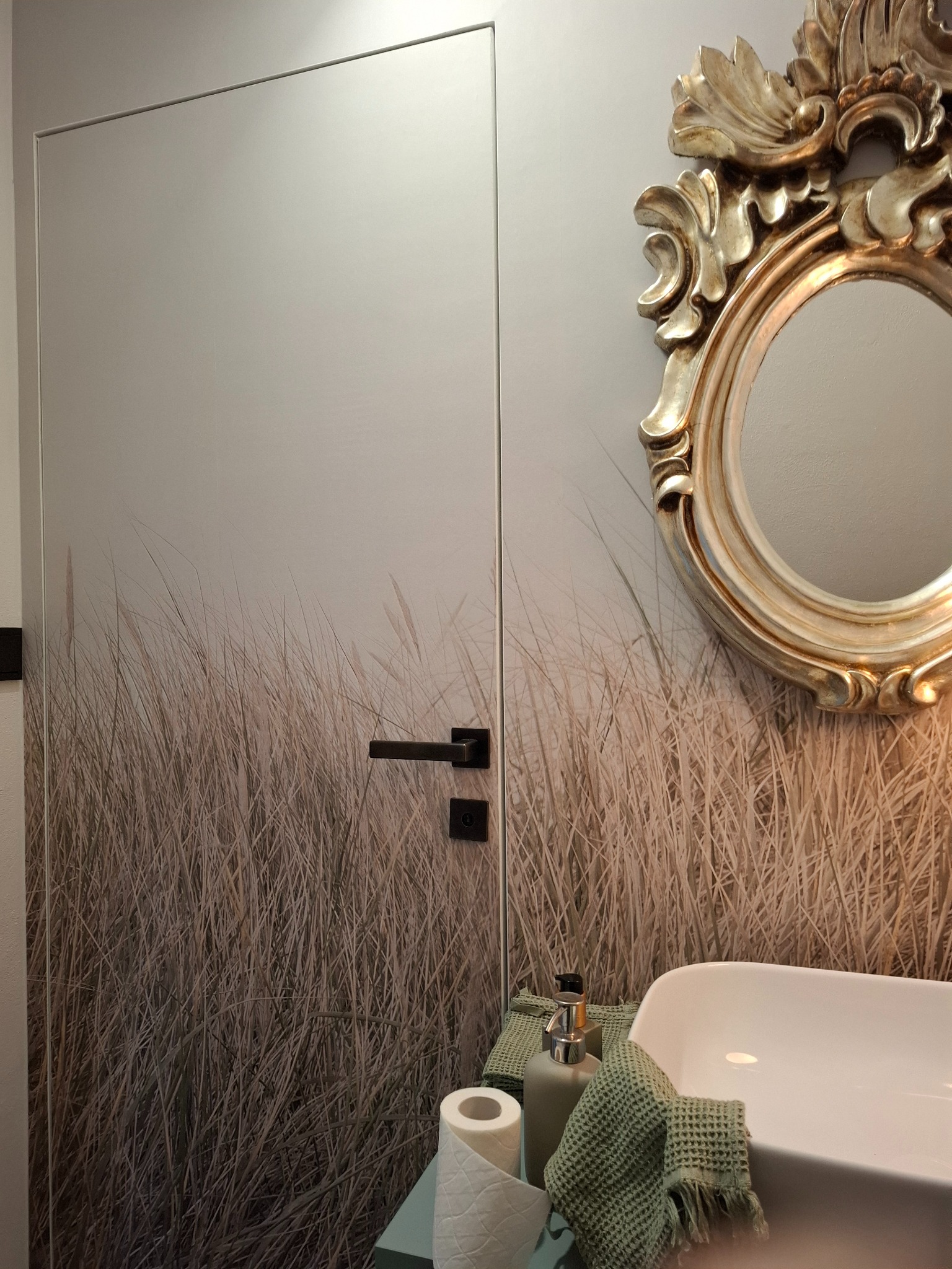 Nowoczesna łazienka z ukrytymi drzwiami z motywem trawy, owalnym lustrem w złotej ramie i białą umywalką z zielonymi akcentami.