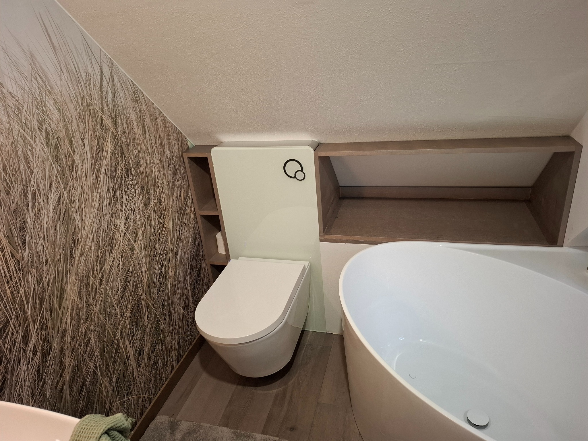 Nowoczesna łazienka na poddaszu: biała wanna owalna, podwieszana toaleta z zabudową, półki z drewnianym wykończeniem, tapeta z motywem trawy. Ukośny sufit.