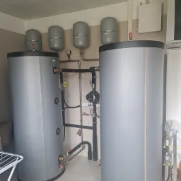 Dwa szare zbiorniki buforowe ciepła z podłączoną instalacją hydrauliczną, z dodatkowymi mniejszymi zbiornikami wyrównawczymi pod sufitem, w pomieszczeniu technicznym z drabinką i suszarką na pranie.