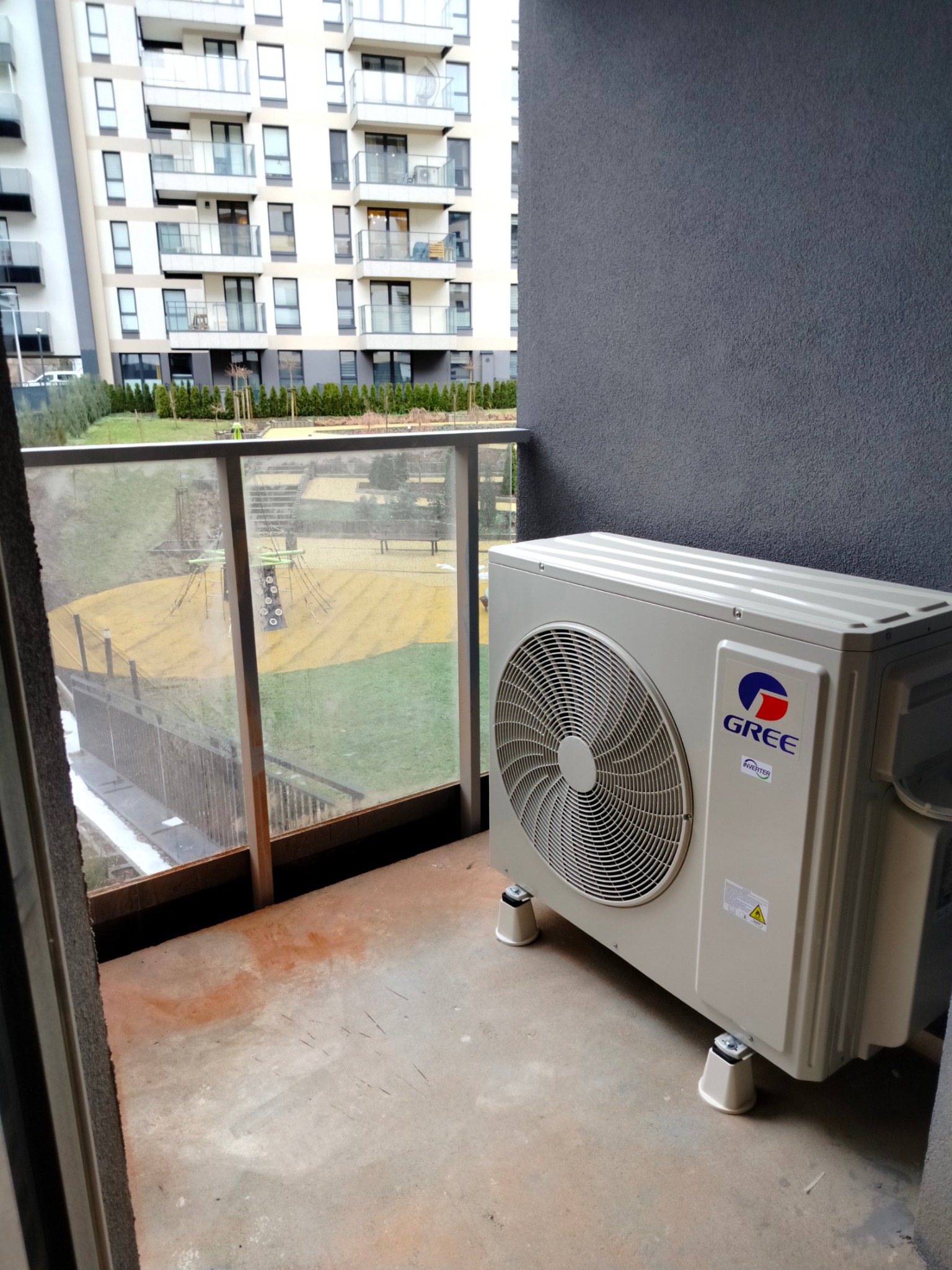 Zewnętrzna jednostka klimatyzacji Gree na balkonie z widokiem na plac zabaw w nowoczesnym budownictwie wielorodzinnym.