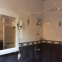 Wykończona łazienka z białymi i ciemnymi marmurkowymi płytkami, lustro z kinkietami i kabina prysznicowa z chromowaną armaturą.