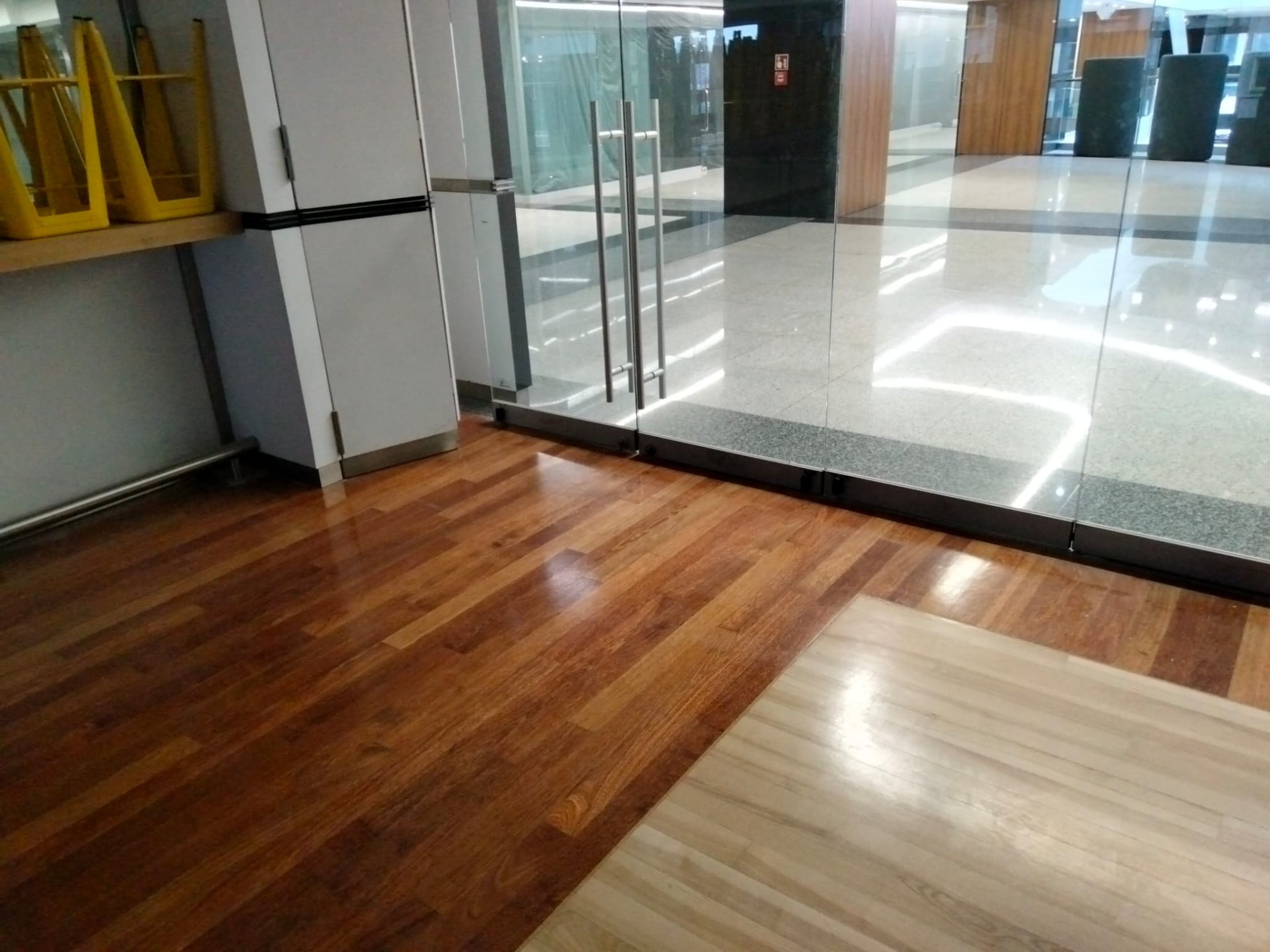 Polerowana drewniana podłoga przechodzi w jaśniejszą, drewnianą powierzchnię, w tle szklane drzwi i błyszcząca, jasna podłoga w budynku.