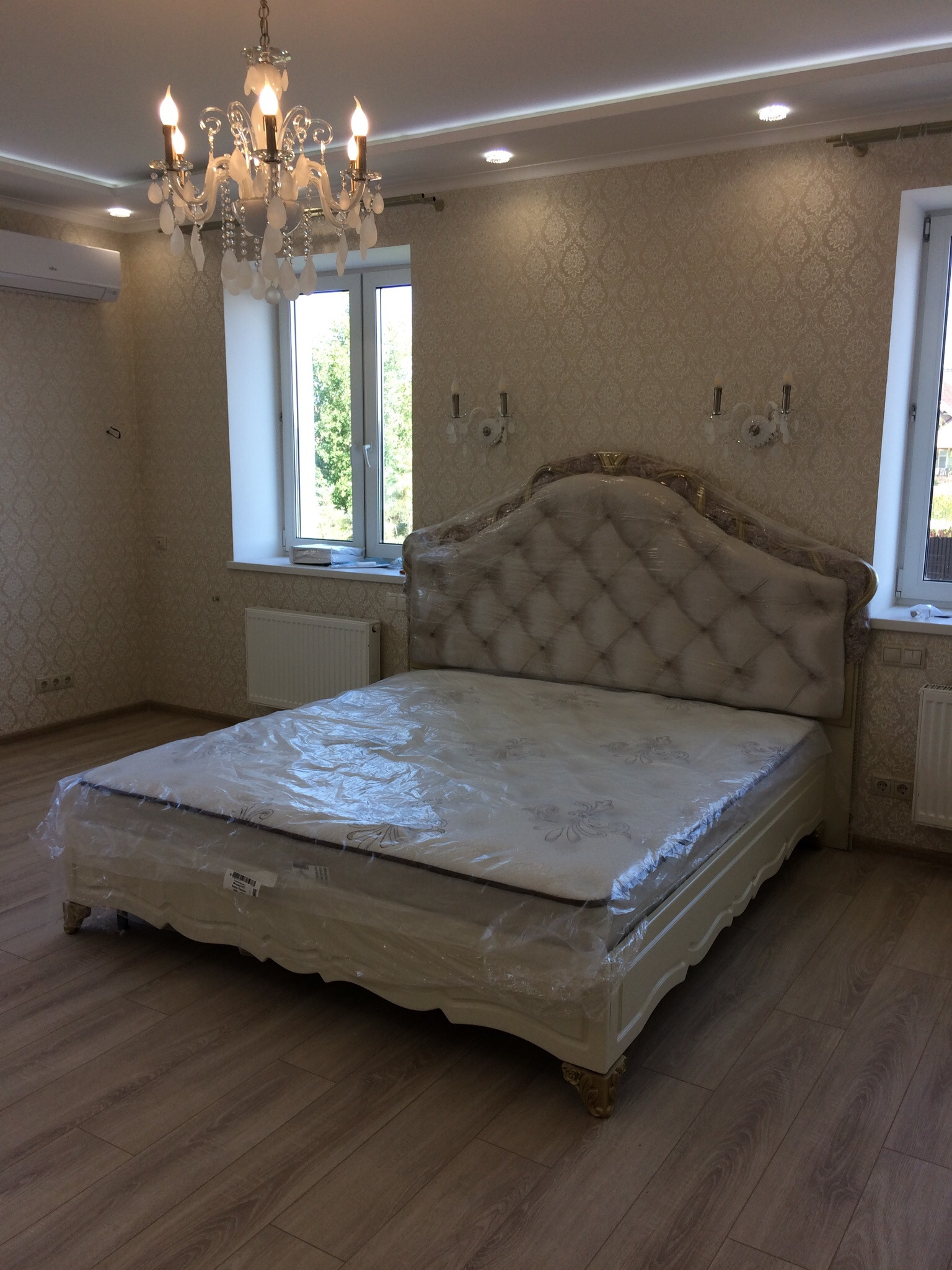 Elegancka sypialnia z łóżkiem w stylu glamour, zabezpieczonym folią ochronną, na świeżo ułożonej podłodze z paneli, z kryształowym żyrandolem i ozdobnymi kinkietami na ścianie w subtelne wzory.