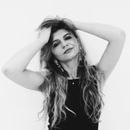 Portret kobiety w czarnej sukience, z rękami uniesionymi nad głową, układającej włosy, z dużymi kolczykami, na jasnym tle, w czerni i bieli.