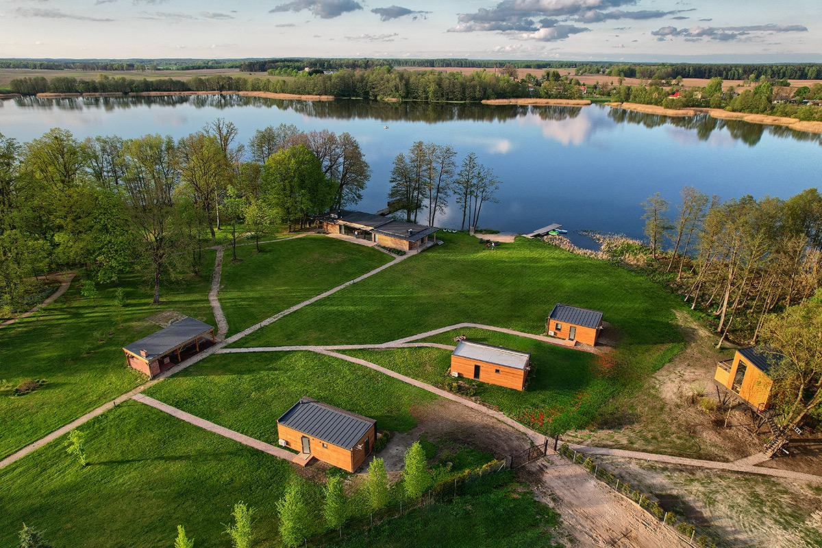 Panoramiczny widok z drona na kompleks drewnianych domków letniskowych nad jeziorem, otoczonych zielenią i ścieżkami. Ujęcie w słoneczny dzień.