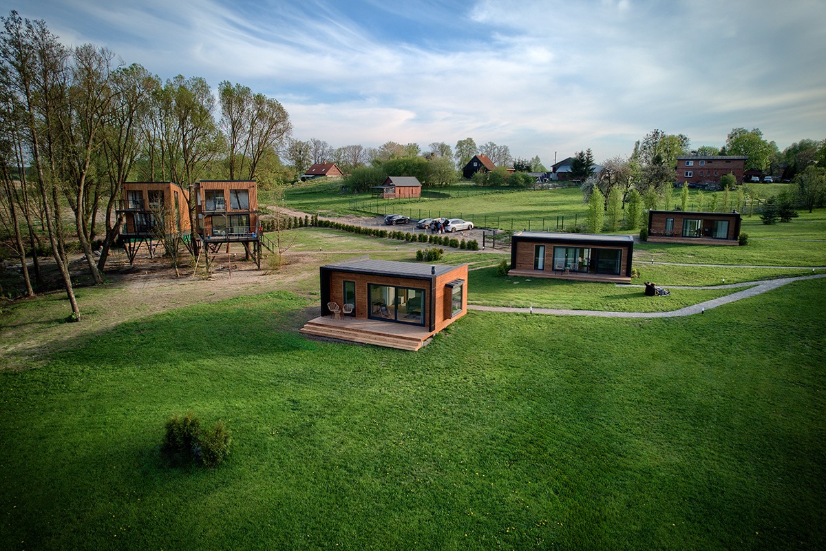 Panoramiczny widok na nowoczesne, drewniane domki letniskowe na zielonym terenie. Domy o minimalistycznym designie, z dużymi przeszkleniami, w otoczeniu drzew i łąk.