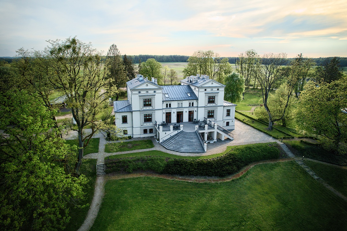 Elewacja dwukondygnacyjnego, białego dworku z szarym dachem, otoczonego zielenią drzew i trawników, sfotografowana z lotu ptaka w słoneczny dzień.