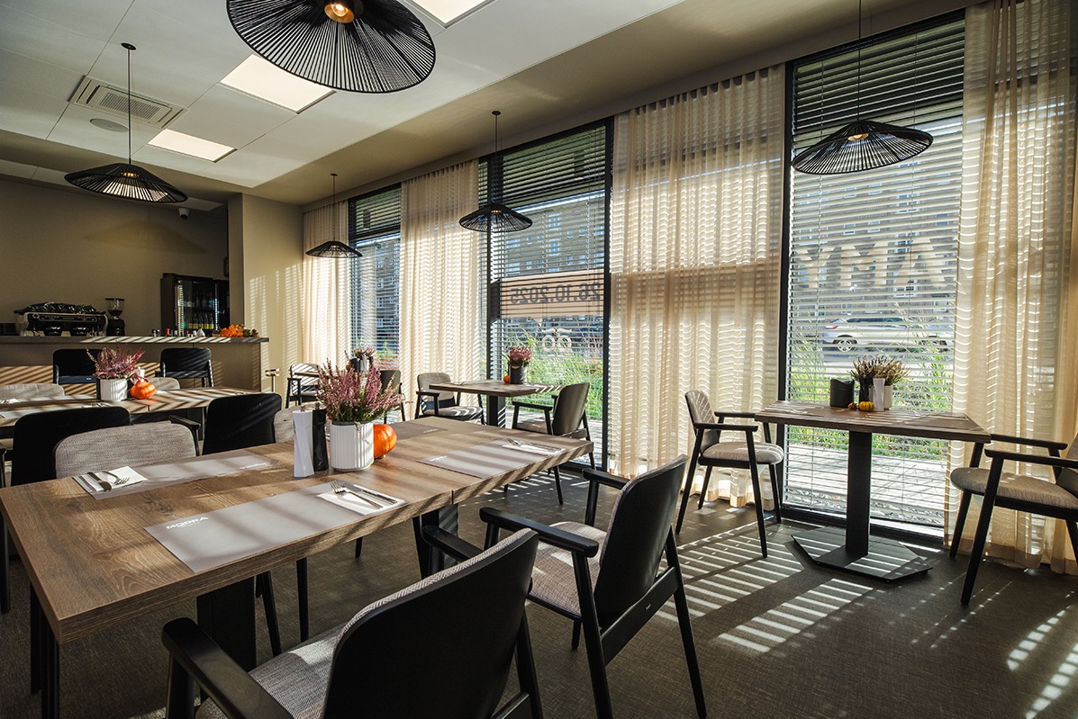 Jasne wnętrze restauracji z drewnianymi stołami, krzesłami i dekoracjami kwiatowymi. Duże okna z żaluzjami wpuszczają naturalne światło. Nowoczesne lampy wiszące.