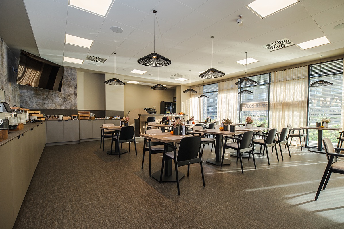 Jasne wnętrze restauracji z drewnianymi stołami, czarnymi krzesłami i designerskimi lampami. Bufet z przekąskami w tle, duże okna z żaluzjami.