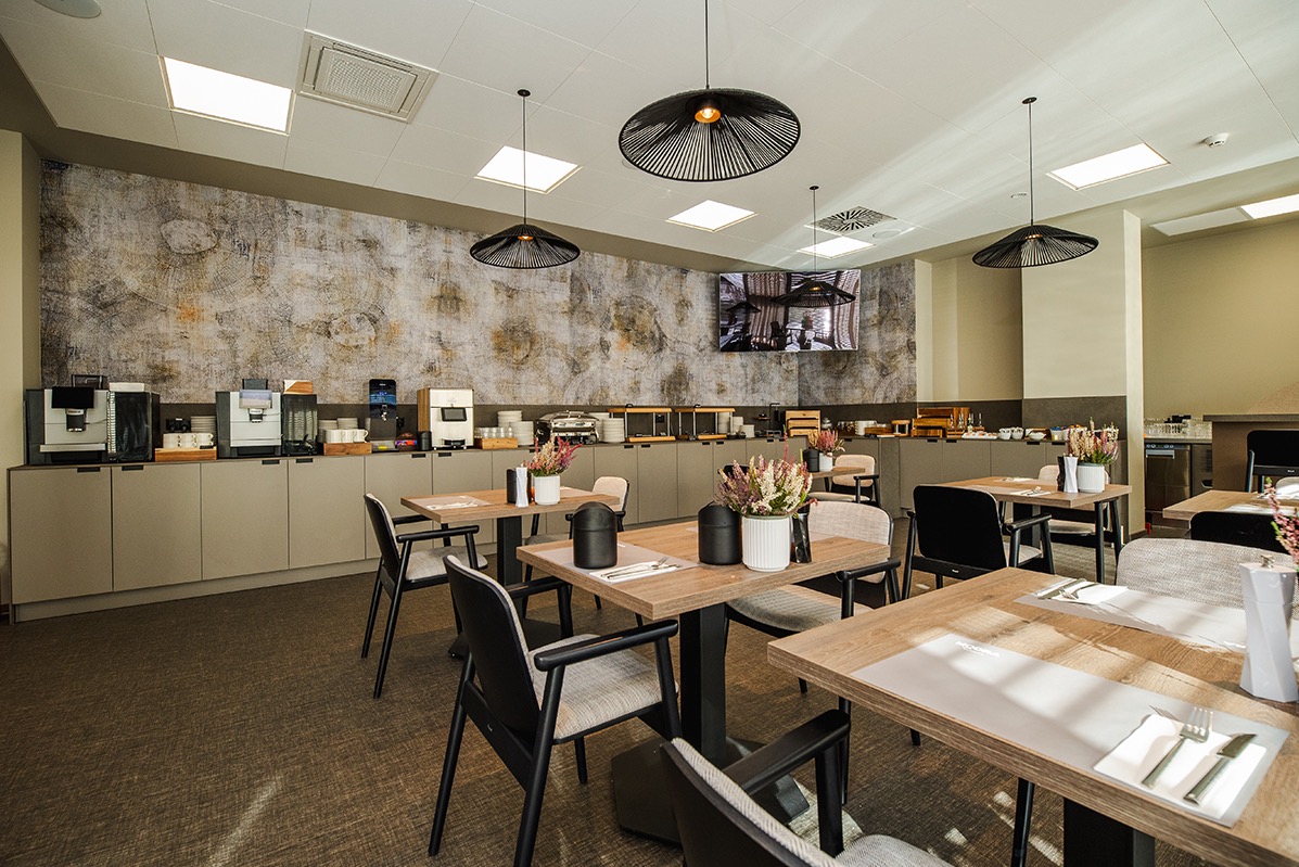 Elegancka sala śniadaniowa hotelu z drewnianymi stolikami, czarnymi krzesłami i bufetem z ekspresami do kawy. Dekoracyjna ściana z motywem drewna i nowoczesne lampy.