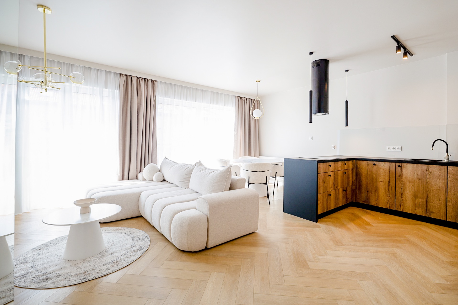 Jasne wnętrze: salon z modułową sofą, okrągłym stolikiem i dywanem, połączony z kuchnią z drewnianymi szafkami. Parkiet w jodełkę, nowoczesne oświetlenie.