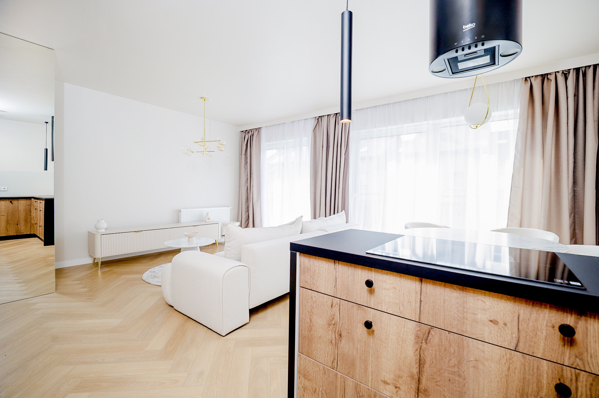 Jasne wnętrze z parkietem w jodełkę, białą sofą i drewnianą zabudową kuchenną z czarnym blatem. Widoczne okno z beżowymi zasłonami i nowoczesne lampy.