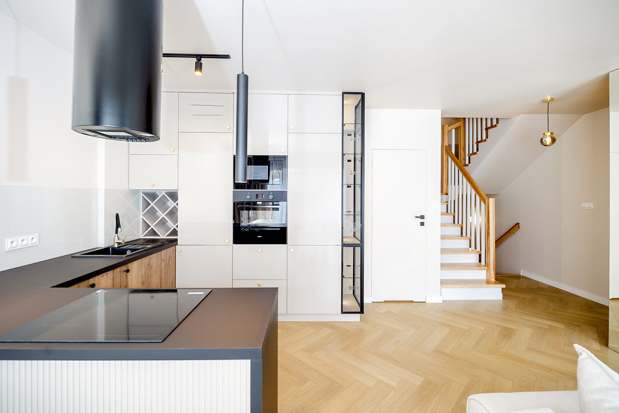 Nowoczesna kuchnia z wyspą i schodami w tle. Białe szafki z czarnymi akcentami, drewniany parkiet w jodełkę. Minimalistyczny design, oświetlenie punktowe.
