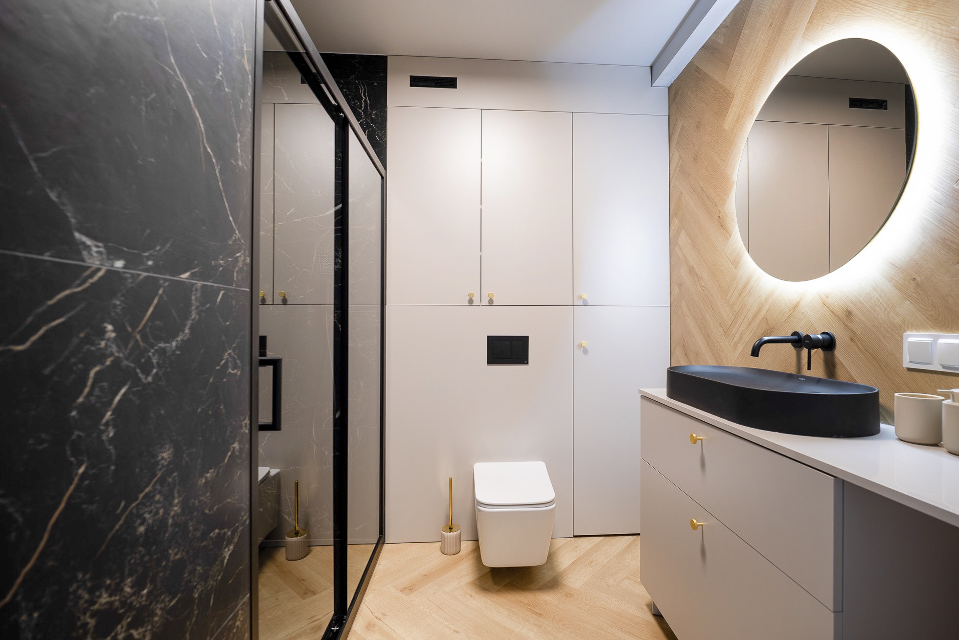 Nowoczesna łazienka z kabiną prysznicową z czarnym marmurem, owalnym lustrem z podświetleniem LED i drewnianą podłogą ułożoną w jodełkę.