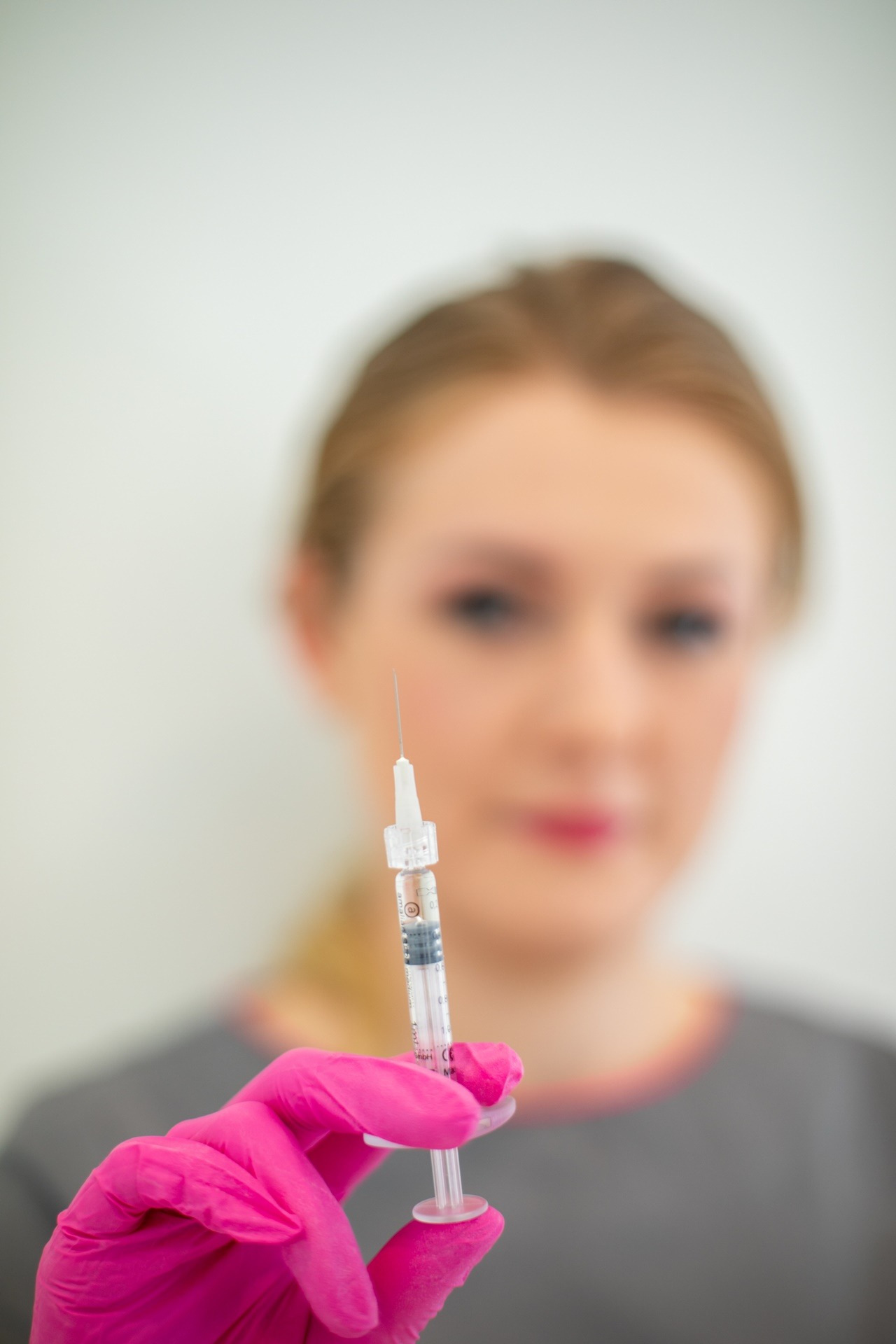 Kobieta w tle, w różowych rękawiczkach trzyma strzykawkę z igłą, zbliżenie na strzykawkę.