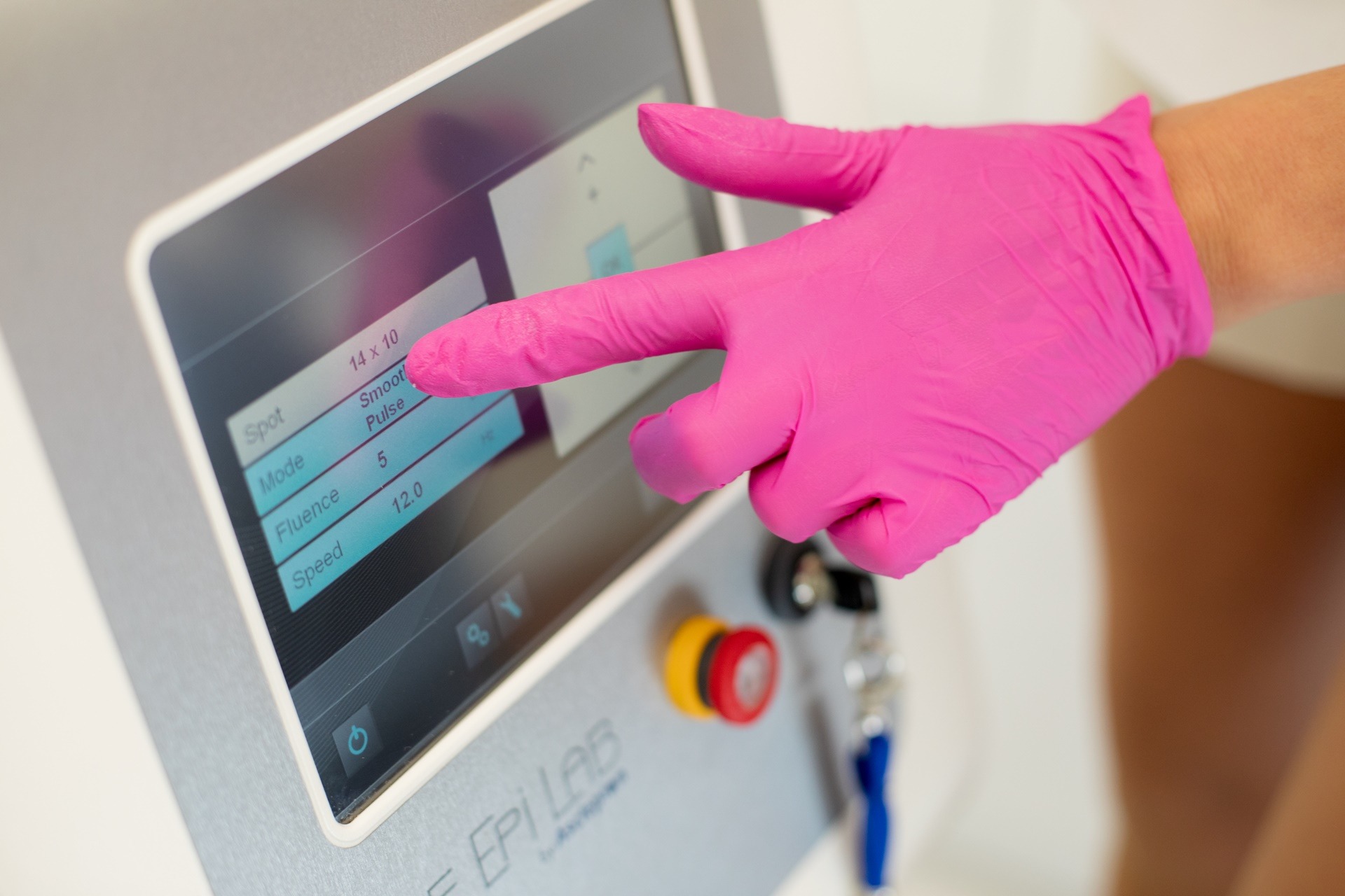 Ręka w różowej rękawiczce dotyka ekranu urządzenia do depilacji laserowej, ustawiając parametry zabiegu.