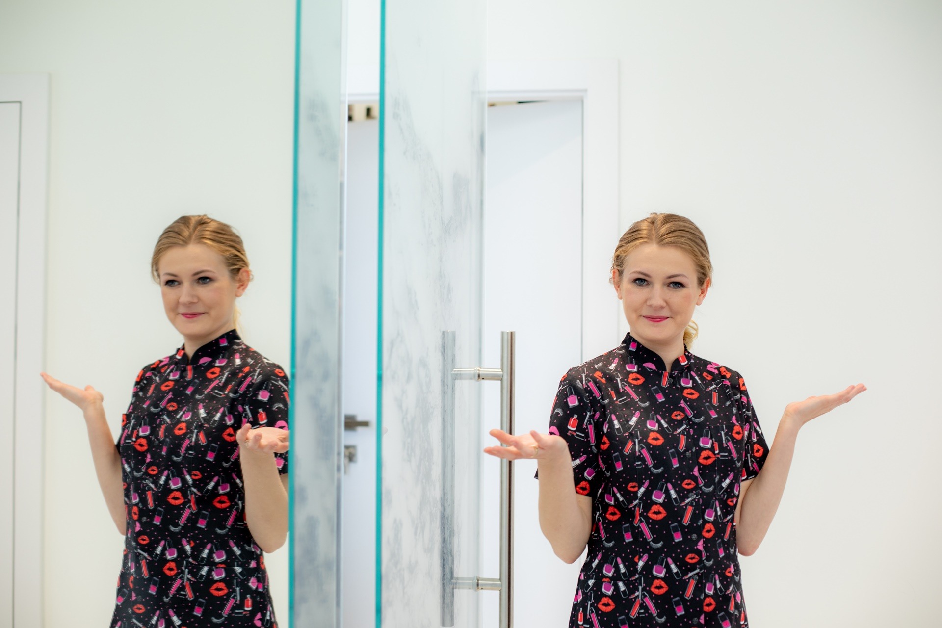 Uśmiechnięta kobieta w uniformie z motywem kosmetyków, stojąca w jasnym wnętrzu z lustrzanymi drzwiami, prezentuje otwartymi dłońmi.