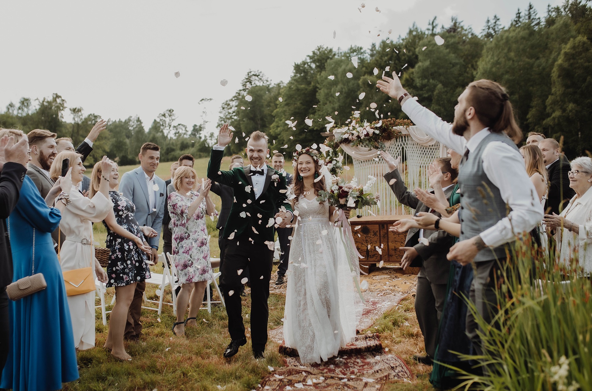 Uśmiechnięta para młoda w otoczeniu gości obsypywana płatkami kwiatów po ceremonii ślubnej na łonie natury; Panna młoda w koronkowej sukni, Pan młody w zielonym aksamitnym smokingu.