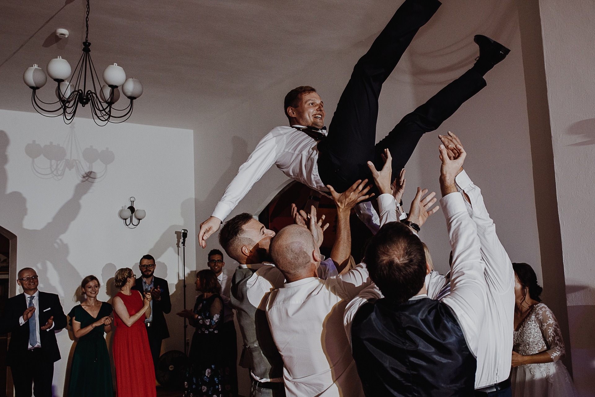 Uśmiechnięty pan młody podrzucany do góry przez gości weselnych w sali z białymi ścianami i żyrandolem, widzowie klaszczą w dłonie.