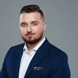 Portret mężczyzny w niebieskiej marynarce i białej koszuli, z poszetką w kieszonce, na szarym tle. Mężczyzna patrzy wprost, z delikatnym uśmiechem.