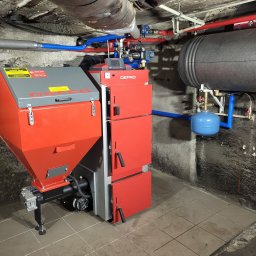 CZECH HYDRO-INSTAL TYMOTEUSZ CZECH - Czerwono-szary piec na paliwo stałe marki DEFRO z podajnikiem, zbiornikiem i sterownikiem, zainstalowany w piwnicy z widocznymi rurami instalacji grzewczej i zbiornikiem akumulacyjnym.