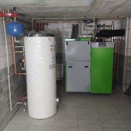 CZECH HYDRO-INSTAL TYMOTEUSZ CZECH - Kotłownia z piecem na paliwo stałe (eko-groszek) w kolorze szaro-zielonym oraz dużym, owiniętym folią ochronną, białym zasobnikiem ciepłej wody użytkowej. Instalacja z rur miedzianych i białych rur...