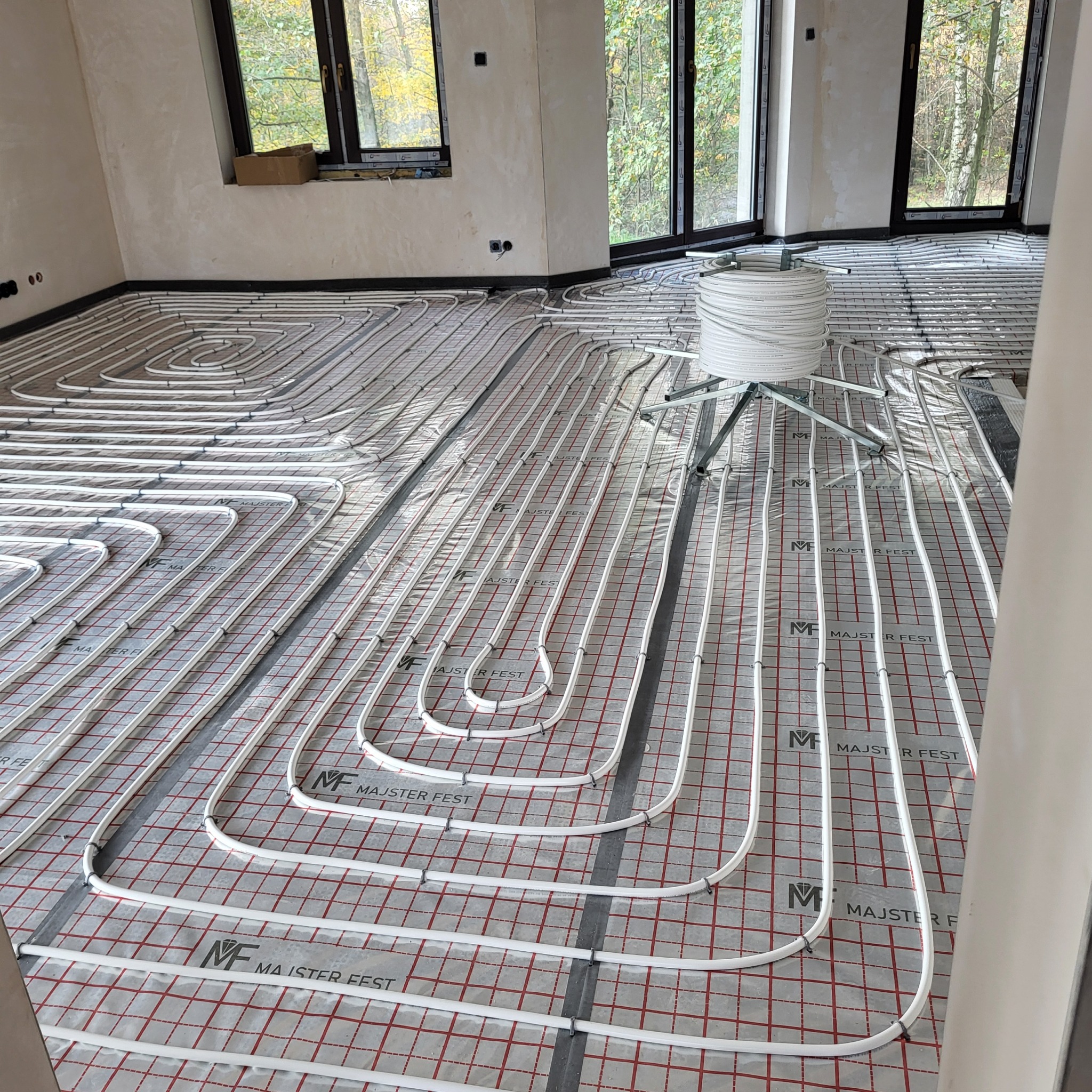 Instalacja ogrzewania podłogowego w nowym domu w Gorzycach, widoczne białe rury ułożone na specjalnej macie z czerwoną kratką, szpula z rurami na stojaku po prawej stronie.